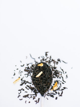 Dark brown black tea leaves blended with dried apply and lemon peel spill from the silver spoon onto the white background