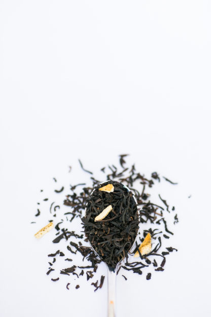 Dark brown black tea leaves blended with dried apply and lemon peel spill from the silver spoon onto the white background