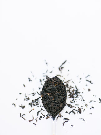 Dark brown black tea leaves overflow the silver spoon onto the white background