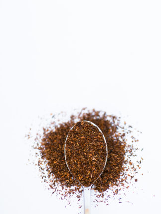 Deep red coarsely ground rooibos needles spill over the silver spoon onto a white background