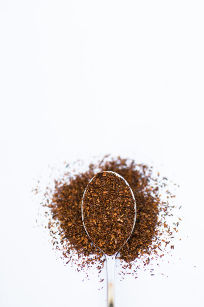 Deep red coarsely ground rooibos needles spill over the silver spoon onto a white background