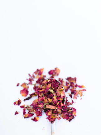 A spectrum of dried red rose petals ranging from dark red to orange red pour out of a silver spoon onto a white background