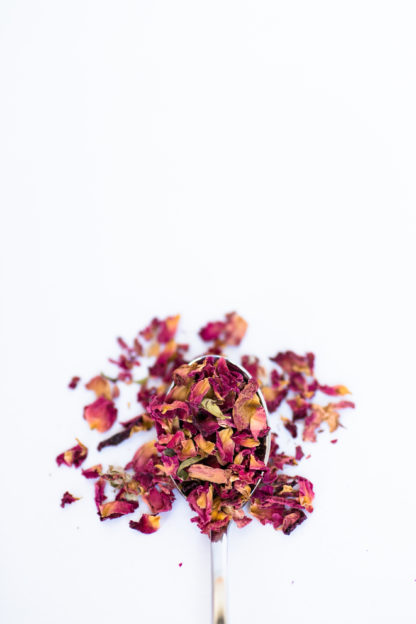 A spectrum of dried red rose petals ranging from dark red to orange red pour out of a silver spoon onto a white background