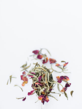 Dark red and orange rose petals blend with light green and white tea needles spilling over the silver spoon onto the white background