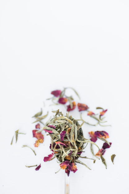 Dark red and orange rose petals blend with light green and white tea needles spilling over the silver spoon onto the white background