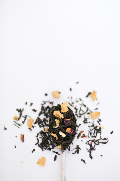 Dark brown black tea leaves blended with lotus flower petals and orange peel cascade over the silver spoon onto the white background