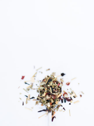 Fluffy green raspberry leaf, red rose hips, light yellow lemon grass and hibiscus flower petals overflow the silver spoon onto a white background