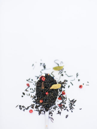 Dark brown black tea leaves, bright red peppercorns, light green cardamom pods and clove pieces overflow the silver spoon onto the white background