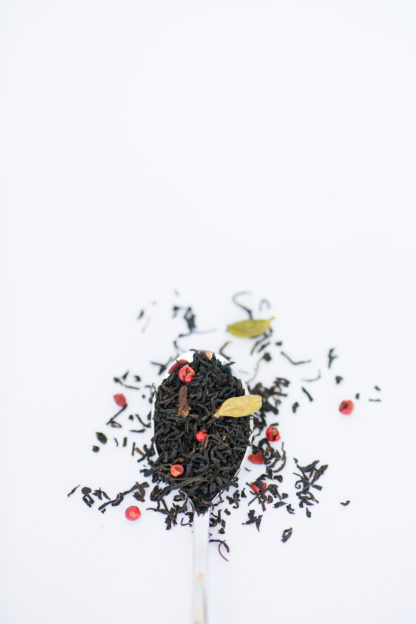 Dark brown black tea leaves, bright red peppercorns, light green cardamom pods and clove pieces overflow the silver spoon onto the white background