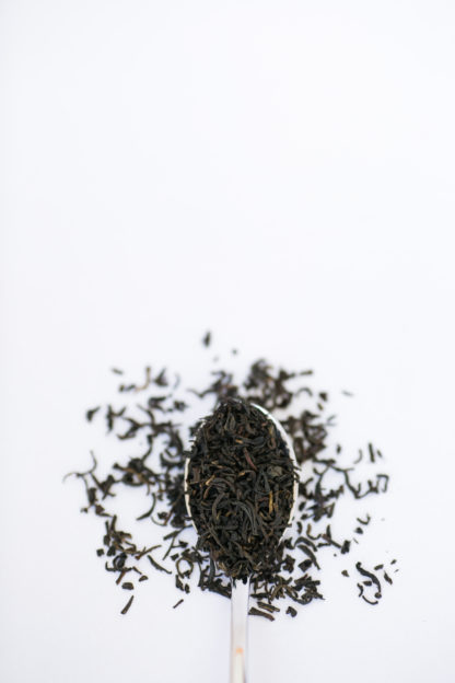 Dark brown black tea leaves overflow the silver spoon onto the white background