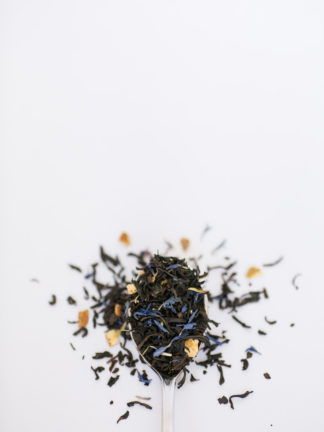 Bright orange peel pieces, corn flowers, and cloves blended with dark brown black tea leaves spilling over the silver spoon onto the white background