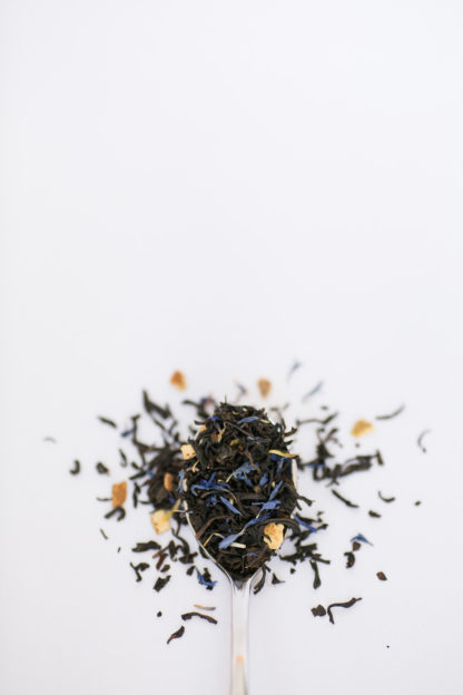 Bright orange peel pieces, corn flowers, and cloves blended with dark brown black tea leaves spilling over the silver spoon onto the white background