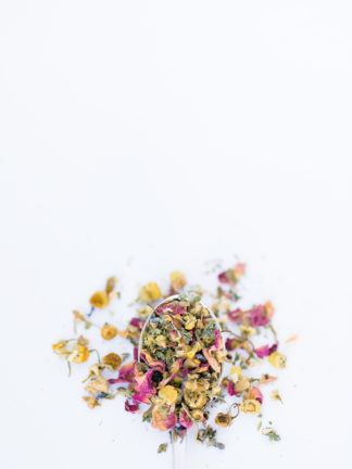 Light yellow chamomile dried florets sprinkled with red rose petals, chopped mint leaves, anise star piedces and seeds flow over the spoon onto a white background