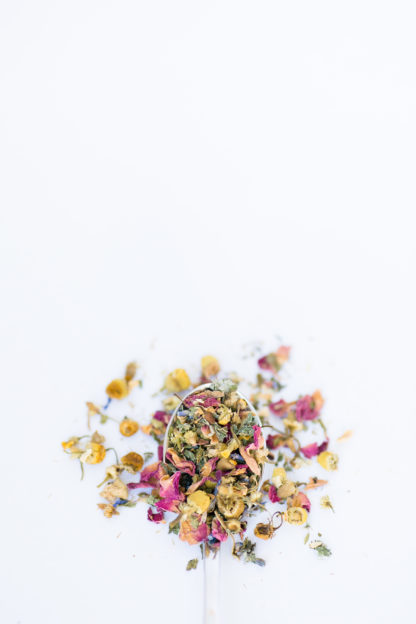 Light yellow chamomile dried florets sprinkled with red rose petals, chopped mint leaves, anise star piedces and seeds flow over the spoon onto a white background