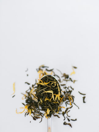Bright yellow marigold flower petas blend with dark green tea leaves flow over the silver spoon onto the white background