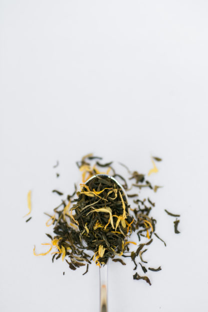 Bright yellow marigold flower petas blend with dark green tea leaves flow over the silver spoon onto the white background