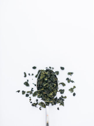 Dark green clumped tea leaves spilling over the silver spoon onto the white background