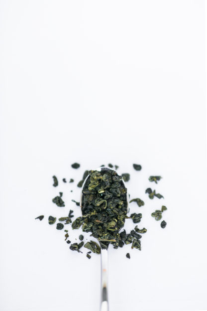 Dark green clumped tea leaves spilling over the silver spoon onto the white background