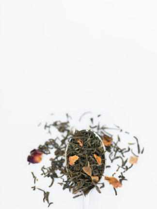 Deep red and organge rose petals are blended into dark green tea leaves spilling over the spoon onto the white background