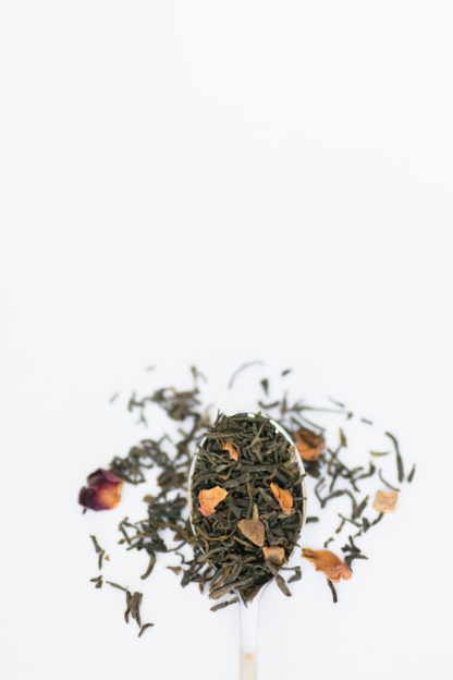 Deep red and organge rose petals are blended into dark green tea leaves spilling over the spoon onto the white background
