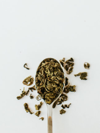 Varigated brown and green classically clumped oolong tea leaves spill over the edge of the silver spoon onto the white background