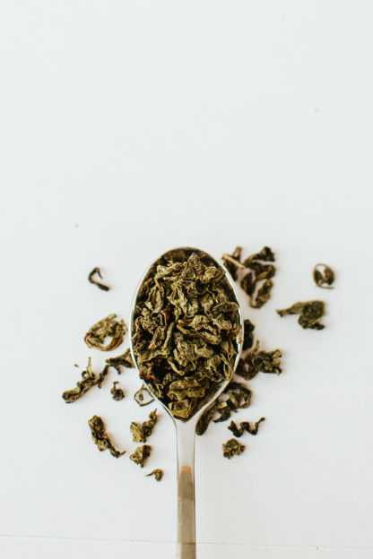 Varigated brown and green classically clumped oolong tea leaves spill over the edge of the silver spoon onto the white background