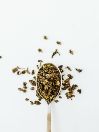 Varigated green and brown classic clumped oolong tea leaves spill over the edge of the silver spoon onto the white background