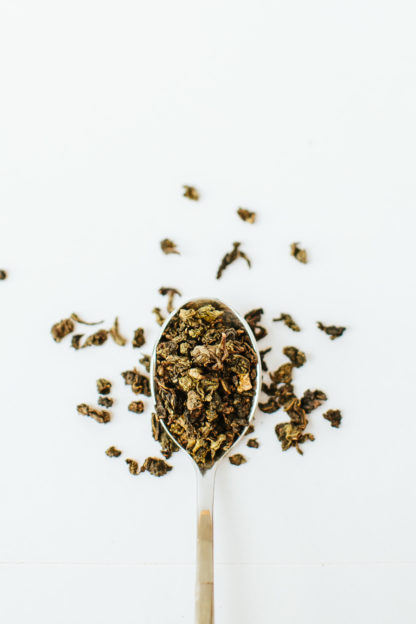 Varigated green and brown classic clumped oolong tea leaves spill over the edge of the silver spoon onto the white background