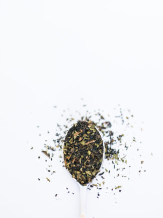 Finely chopped black tea leaves blended with dark green mint pieces overflow the silver spoon onto the white background