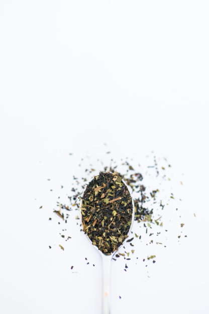 Finely chopped black tea leaves blended with dark green mint pieces overflow the silver spoon onto the white background