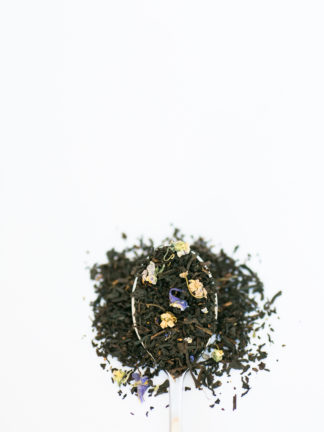 Dark black tea leaves sprinkled with violet flower petals and currents overflow the spoon onto the white background