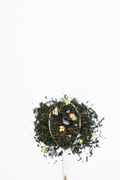 Dark black tea leaves sprinkled with violet flower petals and currents overflow the spoon onto the white background