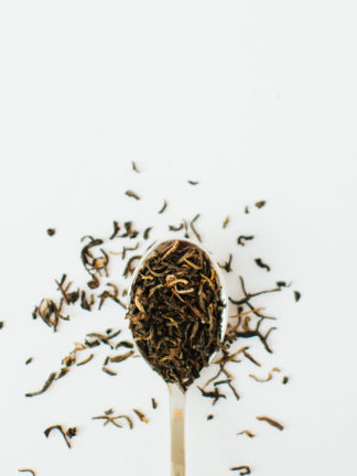 Rich orange tendril-like tea buds blended in a rusty orange black tea in a silver spoon overflowing on a white background