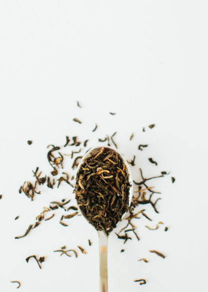 Rich orange tendril-like tea buds blended in a rusty orange black tea in a silver spoon overflowing on a white background