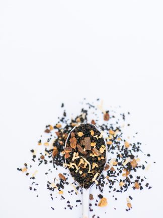 Black tea with white coconut pieces, cardamom, cinnamon bark chips, cloves, and more Middle-Eastern spices spill over the silver spoon onto the white background