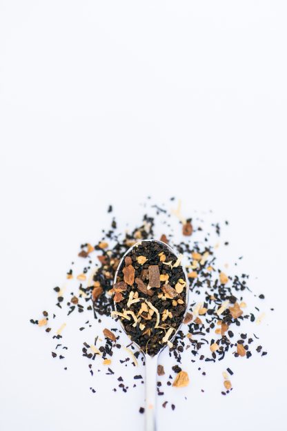 Black tea with white coconut pieces, cardamom, cinnamon bark chips, cloves, and more Middle-Eastern spices spill over the silver spoon onto the white background