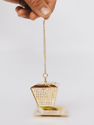 Golden lantern shaped loose leaf tea infuser on chain with cup rim hook suspended above a gold saucer on white background