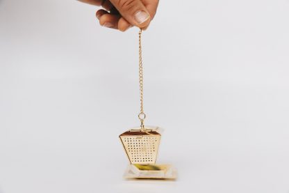 Golden lantern shaped loose leaf tea infuser on chain with cup rim hook suspended above a gold saucer on white background