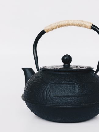 Black cast iron rounded teapot with lotus leaf pattern on surface, short curved spout and heavy duty cast iron arching handle wrapped with tan cord on white background