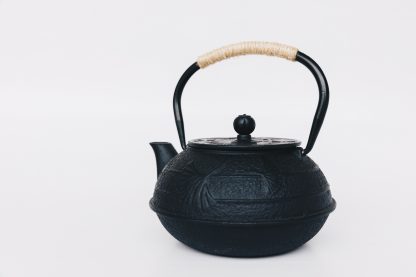 Black cast iron rounded teapot with lotus leaf pattern on surface, short curved spout and heavy duty cast iron arching handle wrapped with tan cord on white background