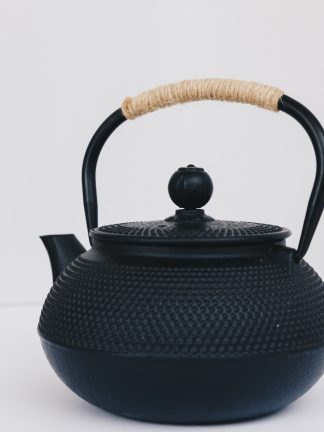 Black cast iron rounded teapot with stippled pattern on surface, short curved spout and heavy duty cast iron arching handle wrapped with tan cord on white background