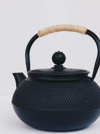 Black cast iron rounded teapot with stippled pattern on surface, short curved spout and heavy duty cast iron arching handle wrapped with tan cord on white background