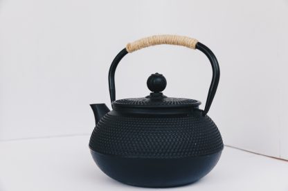 Black cast iron rounded teapot with stippled pattern on surface, short curved spout and heavy duty cast iron arching handle wrapped with tan cord on white background