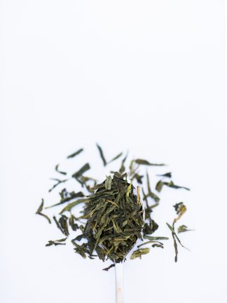 Flat dried green tea leaves overflow a silver spoon onto a white background