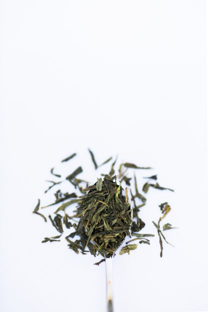 Flat dried green tea leaves overflow a silver spoon onto a white background