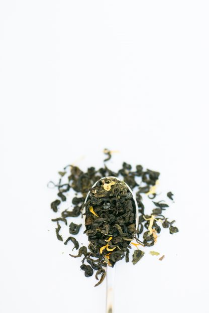 Dark green clumped loose leaf tea leaves mixed with safflowers and mint cascading over a silver spoon on a white background