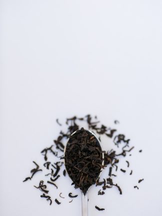 Small dark black tea leaves cascade from a silver spoon onto a white background