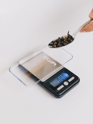 Small loose leaf tea scale with clear lid and tray for weighing teas to the .01 gram with silver tea spoon containing tea to be weighed on white background