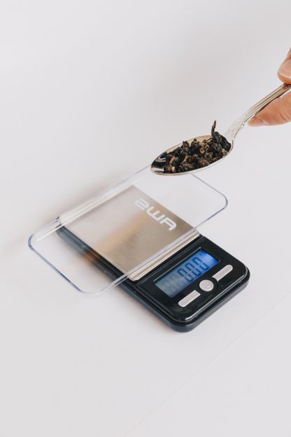 Small loose leaf tea scale with clear lid and tray for weighing teas to the .01 gram with silver tea spoon containing tea to be weighed on white background