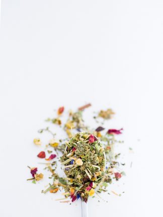 Beautiful dried chamomile florets blended with rose petals, lemon peal, lemon verbena, lemon grass overflow a silver spoon onto a white background